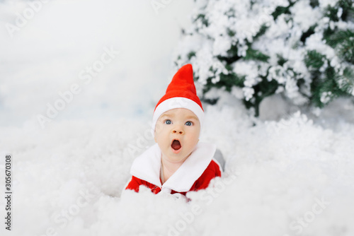 A baby in a little Santa costume and hat lies on its back in the artificial snow. The kid is surprised by the sale. The concept of Christmas holidays. Greeting card © Sunshine
