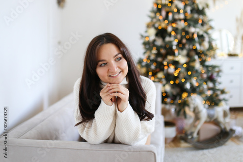 The concept of new year's shooting. Beautiful attractive brunette girl in a white cozy warm dress lying and fooling around on the floor upside down near the Christmas tree gifts and dresser.