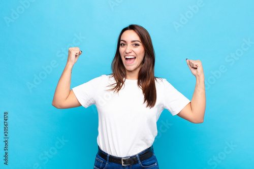 young pretty woman feeling happy, satisfied and powerful, flexing fit and muscular biceps, looking strong after the gym against blue wall