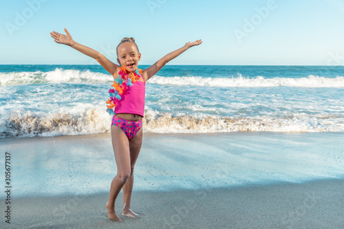 A cute little smiling girl in a colorful pink swimsuit with hawaiian floral garland on the beach, open arms embracing nature, copy space