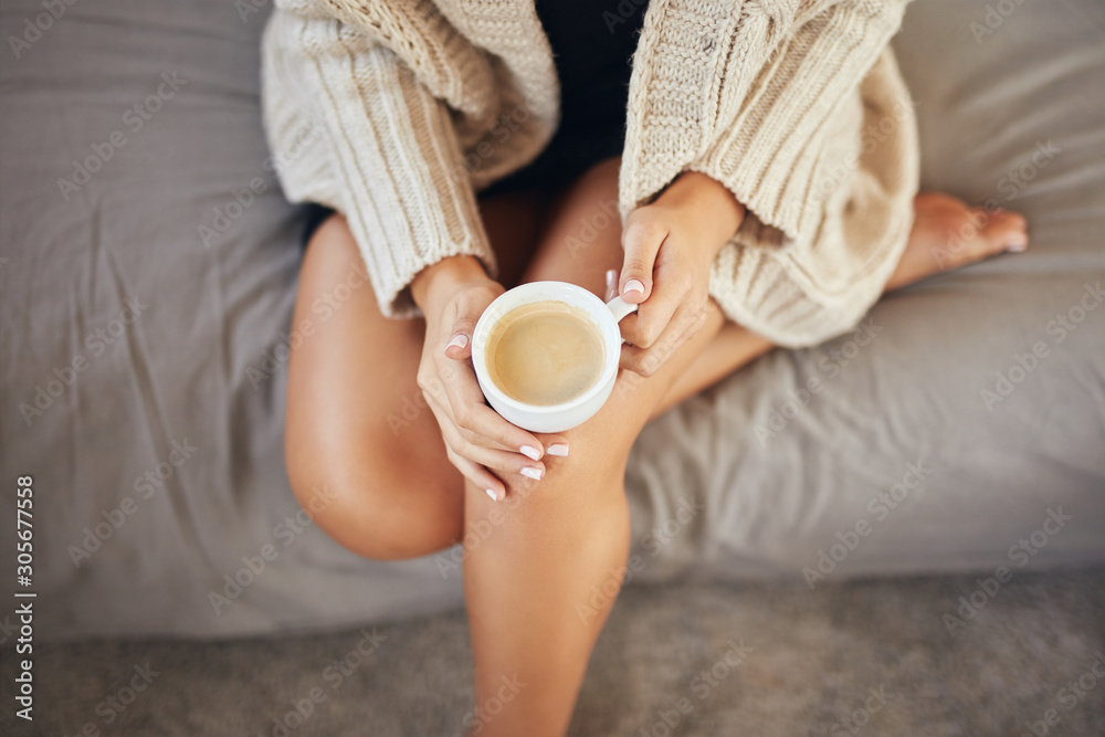 Top view of caucasian woman dressed in beige sweater sitting on bed and holding fresh morning coffee.
