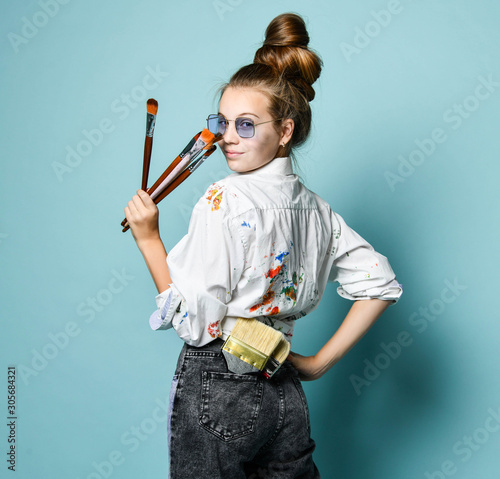 Young artist woman with a set of brushes in shirt with paint stains is at work, standing back, looking over her shoulder photo