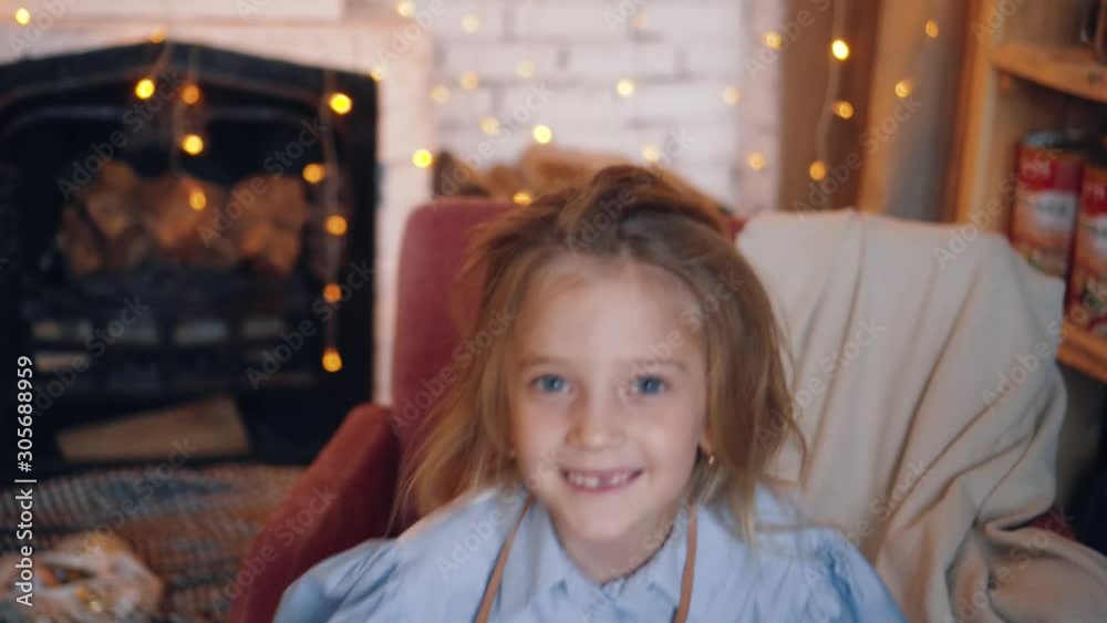 Little girl plays the fool, she is making fun with closing her face and opening it again. Playing child at new year eve. Garlands and light on the bokeh.