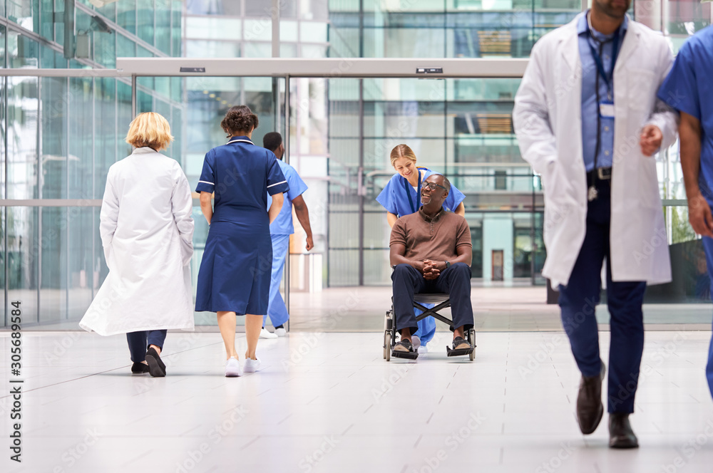 Female Nurse Wearing Scrubs Wheeling Patient In Wheelchair Through Lobby Of Modern Hospital Building Female Nurse Wearing Scrubs Wheeling Patient In Wheelchair Through Lobby Of Modern Hospital Building