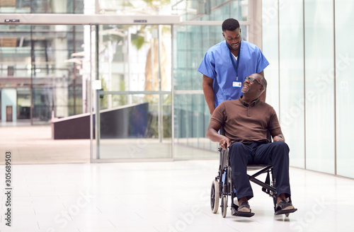 Schilderij op canvas Male Nurse Wearing Scrubs Wheeling Patient In Wheelchair Through Lobby Of Modern