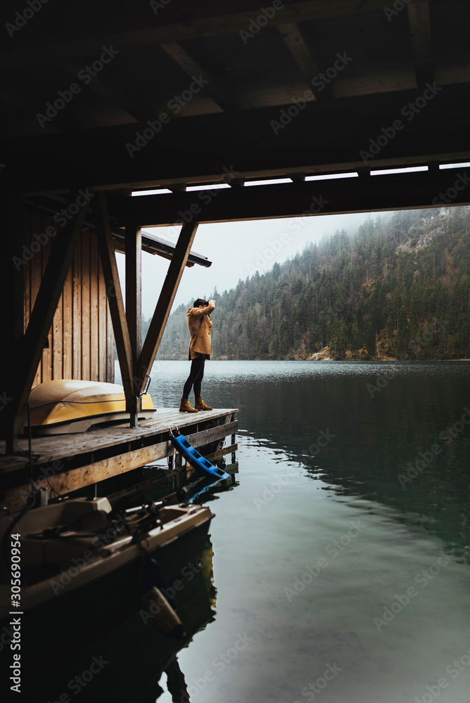 Full body shot of young male photographer in brown coat taking pictures with a small mirrorless camera standing on a lake house deck in lake plansee, Austria