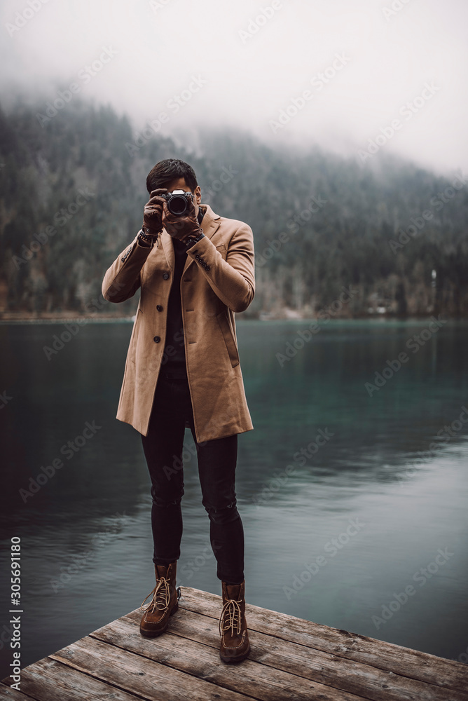 Full body shot of young male photographer in brown coat taking pictures with a small mirrorless camera standing on a lake house deck in lake plansee Austria Stock Adobe Stock