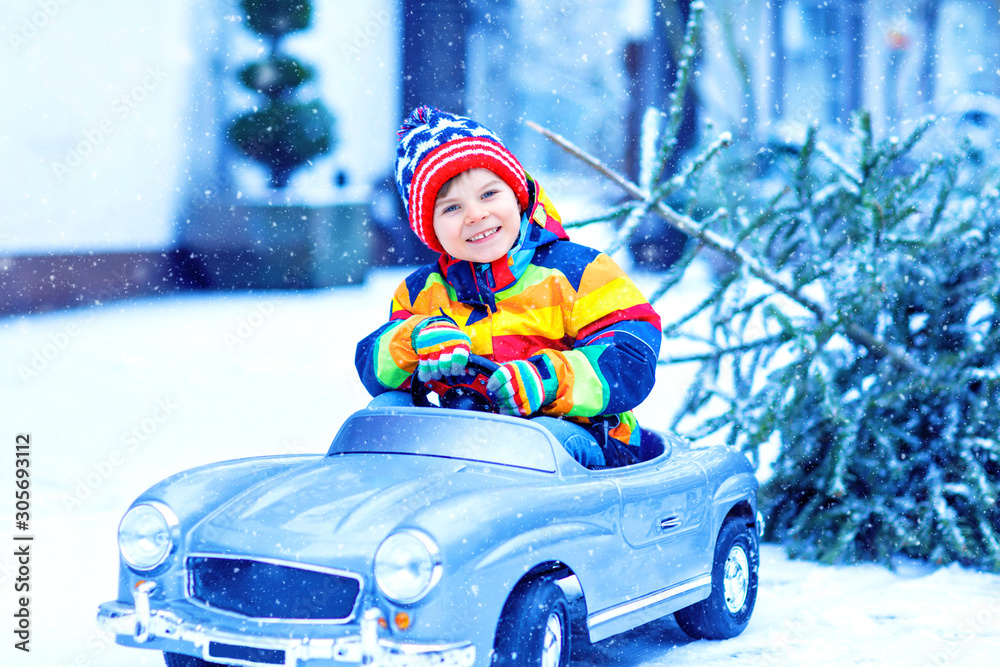 Beautiful little smiling kid boy driving toy car with Christmas tree. Happy child in winter fashion clothes bringing hewed xmas tree from snowy forest. Family, tradition, holiday.
