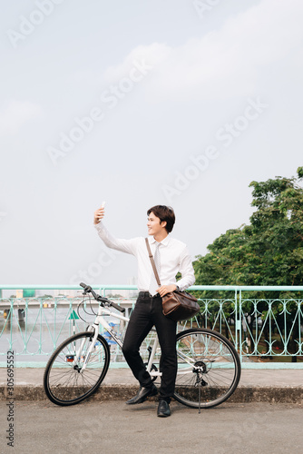 Fototapeta Naklejka Na Ścianę i Meble -  Young man taking a photo with his smart phone while standing on the city street with his bicycle beside him