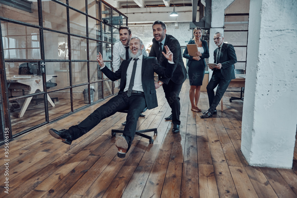 Obraz premium Playful businessmen pushing their boss on the office chair while running in the hallway
