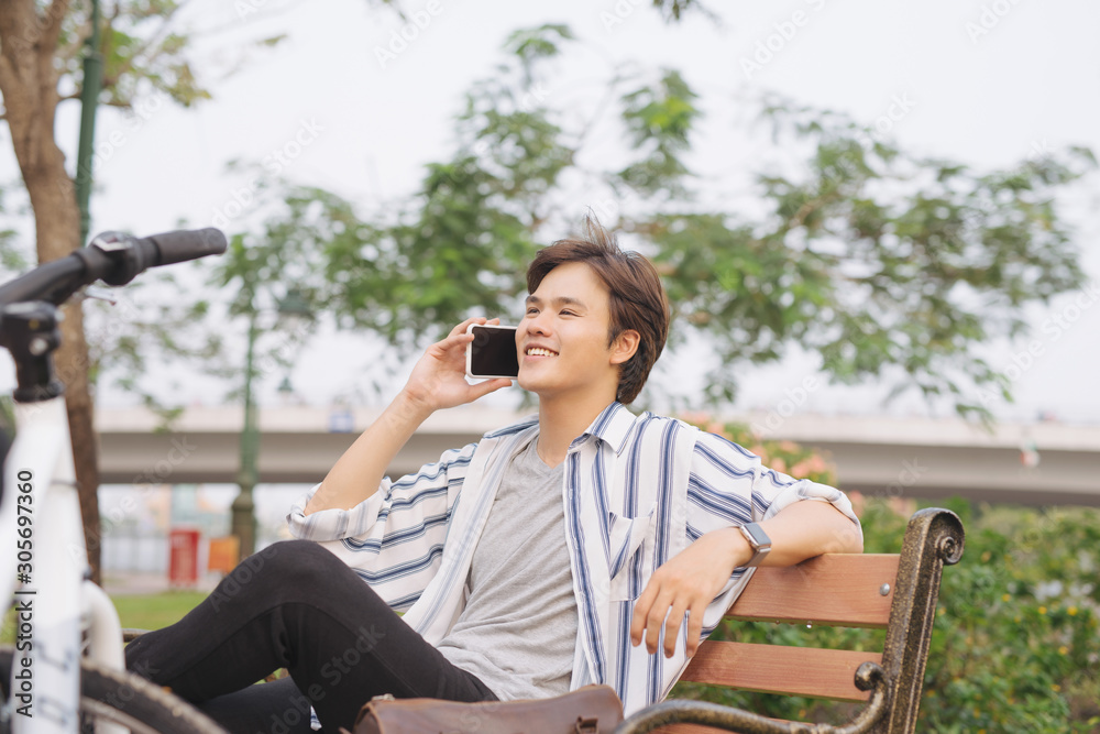Young handsome businessman talking by mobile phone while sitting on bench in park after riding bicycle
