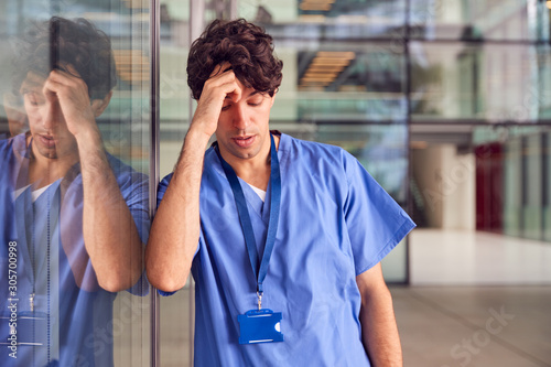Tired Young Male Doctor Wearing Scrubs Leaning Against Wall In Modern Hospital Building photo