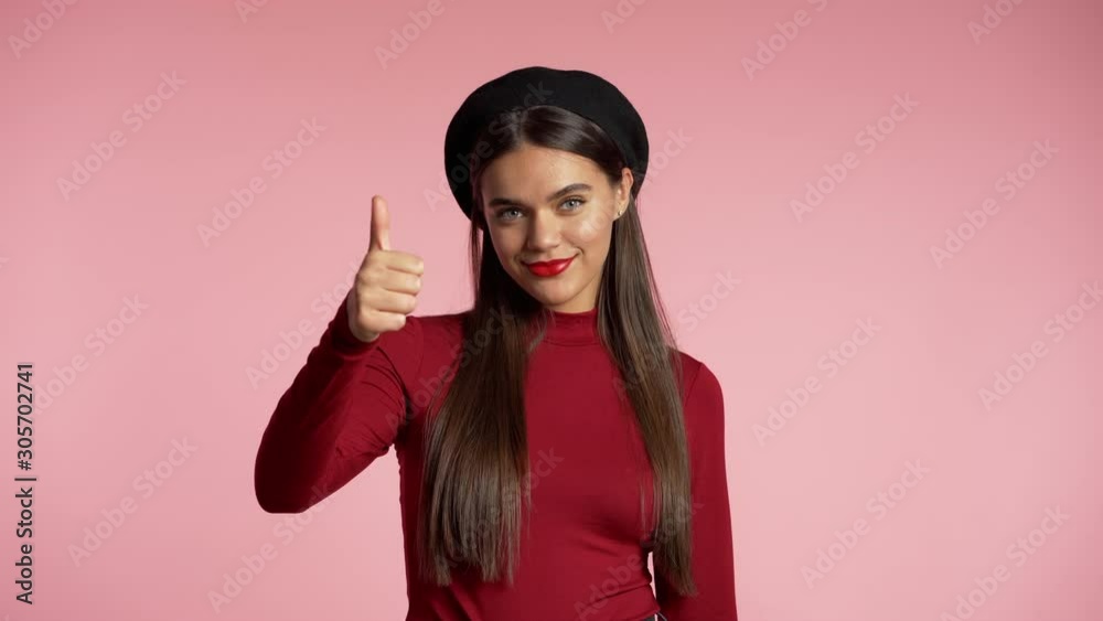 Winner. Success. Positive girl making thumbs up sign over pink background and smiles to camera. Body language. Young hipster woman with perfect red lips make-up and french hat.