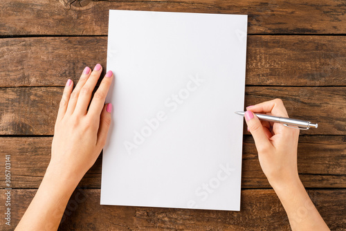 Woman’s hands writing with pen over blank white paper sheet on wooden table. Top view photo