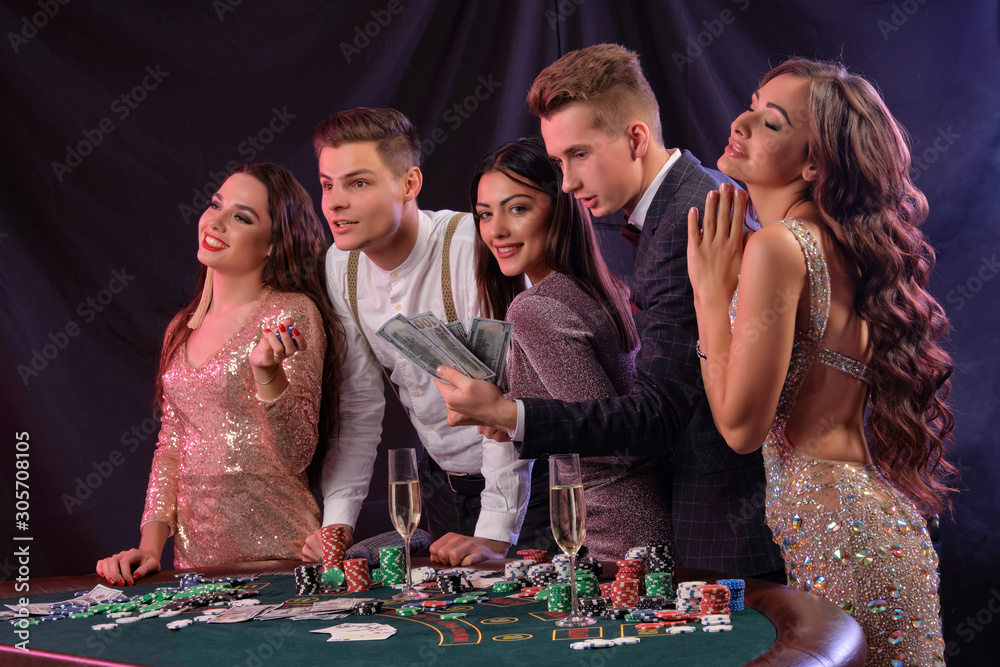 Friends playing poker at casino, at table with stacks of chips, money, cards, champagne on it. Celebrating win, smiling. Black background. Close-up. Friends playing poker at casino, at table with stacks of chips, money, cards, champagne on it. Celebrating win, smiling. Black background. Close-up.