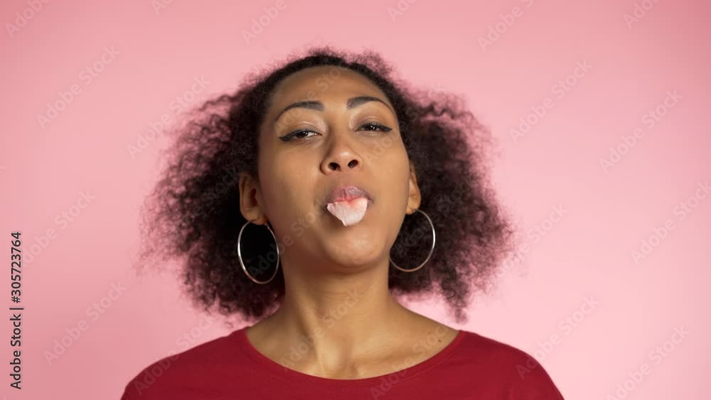 Playful girl in red wear blows bubblegum, chewing gum in slow motion. Pretty african american woman stands on pink background. Playful girl in red wear blows bubblegum, chewing gum in slow motion. Pretty african american woman stands on pink background.