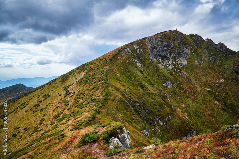 Fototapeta premium Hiking path in green mountains peaks and hills with yellow grass outdoor view, autumn rural landscape with cloudy sky. Adventure mountain trekking concept. Moody light. Panoramic view background