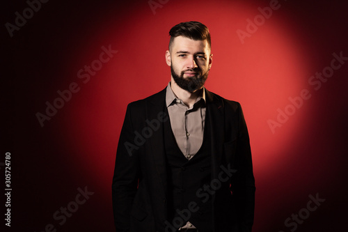 Young serious bearded man in black suit on red background. A man looks into the camera