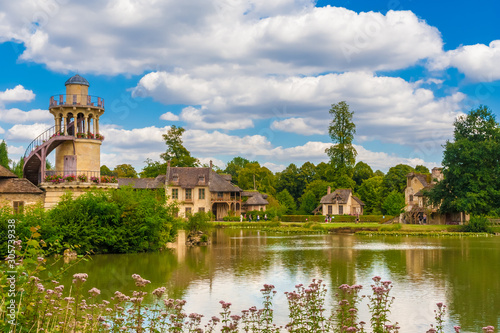 Beautiful panoramic landscape view of the Marlborough Tower, the Queen’s House, the boudoir and the mill from the lake in the Queen’s Hamlet at the Petit Trianon park of Versailles on a summer day.