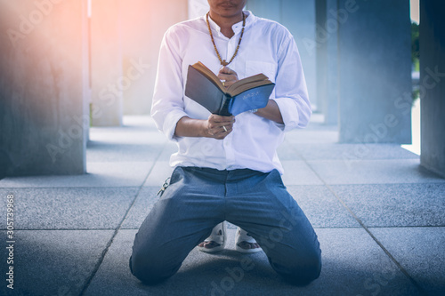 Handsome young Asian wearing his rosary crucifix symbol. He knelt and read the holy bible expresses the wish of God. Sunlight Ladcheag concrete walkway. Concept of Christian Prayer. photo