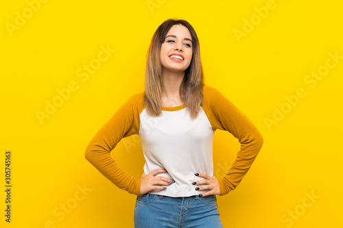 Pretty young woman over isolated yellow wall posing with arms at hip and smiling