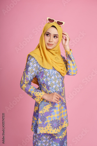 Beautiful female model wearing batik design "baju kurung" with yellow colored hijab, sitting on a chair isolated over pink background. Eidul fitri fashion and beauty concept.