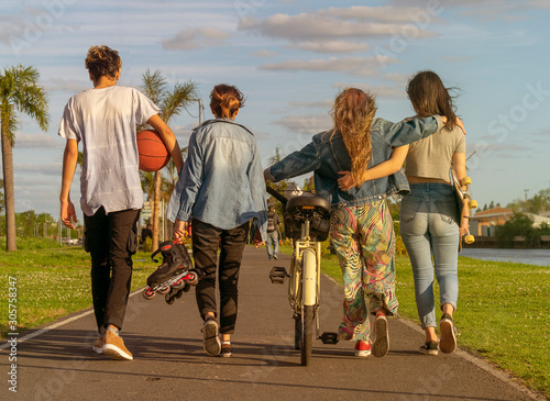 Portrait from behind of four teenagers walking along a path in a park with a bike, a skateboard, roller skates and a basketball #305758347