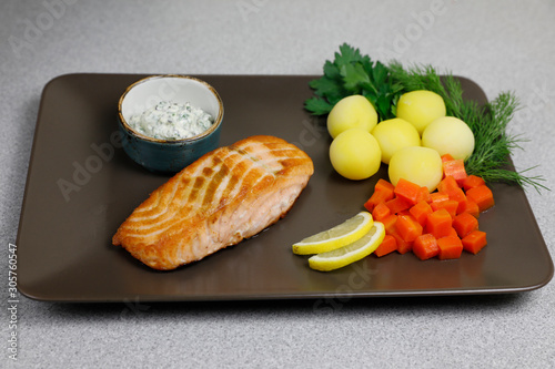 Grilled salmon on a brown rectangular plate with vegetables, potatoes, carrot, lemon, and greens: parsley and dill; with sauce dip. Top view