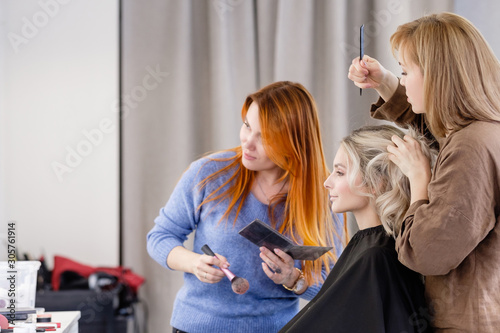 Women redhear make-up artist and hairdresser do hairstyle and stylish makeup for beautiful young blonde girl before start of fashion show. Colorful makeup design beauty and art salon concept