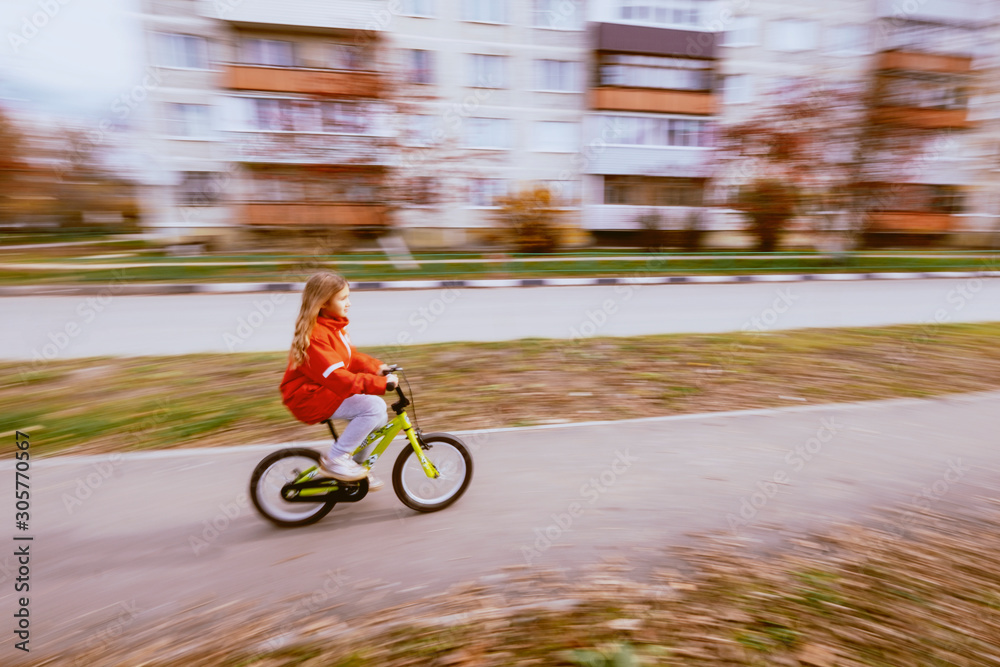 Young blonde girl in red jacket riding bike. Child bicycle in city. Little lady enjoying bike ride on her way to school or park. Sport for kids and eco transport concept.