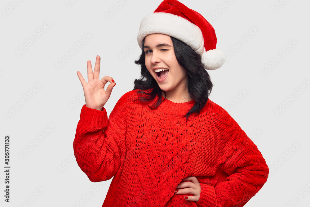 Excited brunette girl in a red sweater and Santa hat showing OK gesture and winking at the camera Excited brunette girl in a red sweater and Santa hat showing OK gesture and winking at the camera