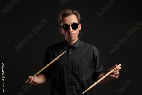 Stylish man drummer in a black shirt and sun glasses playing drums with sticks over dark background.