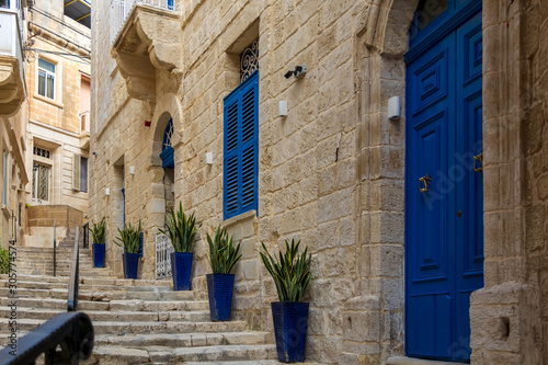 Narrow charming street in Birgu, Malta, with limestone medieval buildings with navy blue doors and window shutters and plants in blue pots along the walls.