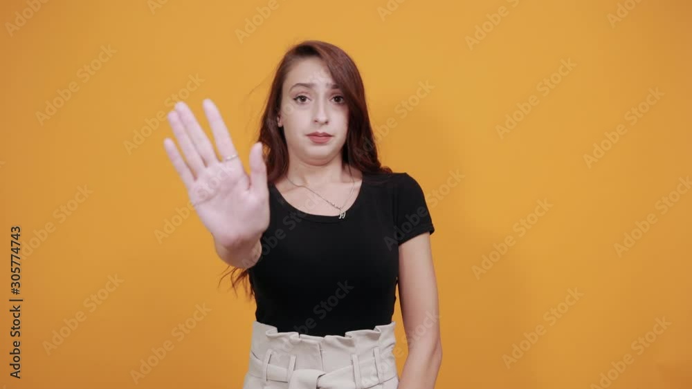 Serious young woman in black shirt, doing stop NO gesture, disappointed in all isolated on orange background in studio. People sincere emotions, health concept.