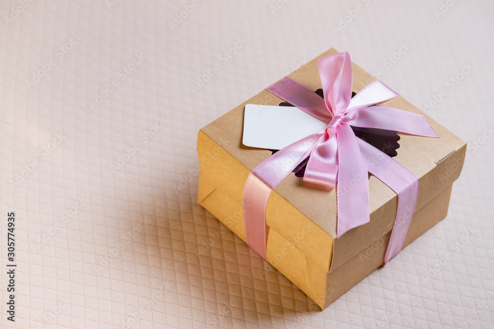 A square gift box with a pink ribbon on a light pink background. Cupcakes inside. Space for text, empty note tied over. A square gift box with a pink ribbon on a light pink background. Cupcakes inside. Space for text, empty note tied over.