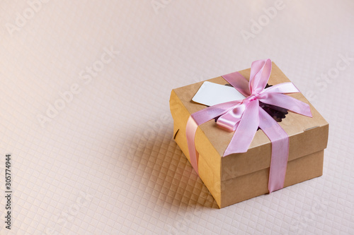 A square gift box with a pink ribbon on a light pink background. Cupcakes inside. Space for text, empty note tied over.