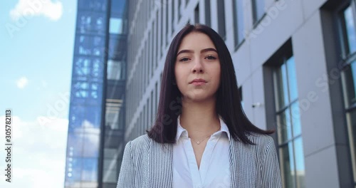 Portrait of the young attractive Caucasian brunette woman in the shirt and jacket looking straight at the camra in the big modern building background. Outdoor. photo