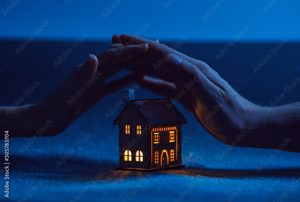 home protection. male and female hand on a small house in which the windows glow with warm light - obrazy, fototapety, plakaty 