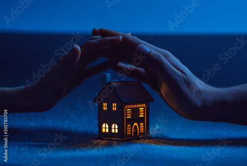 home protection. male and female hand on a small house in which the windows glow with warm light