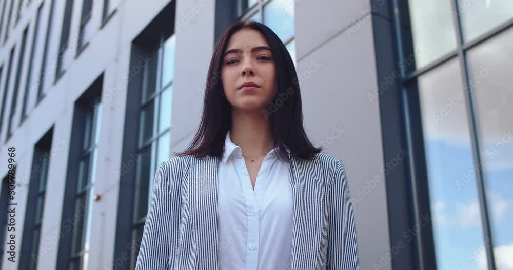 Portrait shot of the young Caucasian attractive businesswoman with long dark hair standing at the office building, looking at the side and then urning face to the camera. Close up. Outdoors. Portrait shot of the young Caucasian attractive businesswoman with long dark hair standing at the office building, looking at the side and then urning face to the camera. Close up. Outdoors.