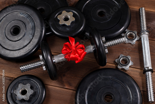gift - dumbbells tied with a red ribbon on a dark wooden table along with sports equipment