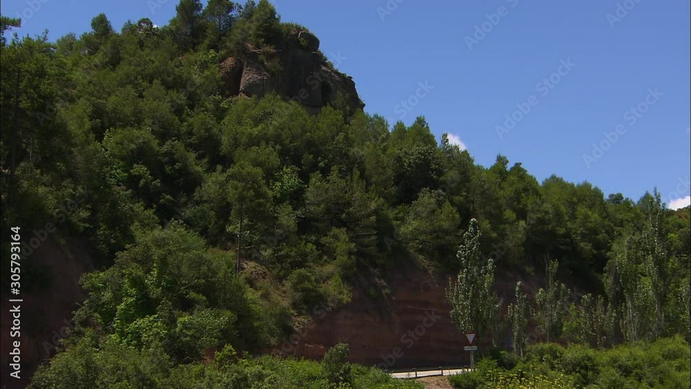 Wide low-angle still shot of a steep Montserrat mountain with pine trees, and a road passing at its base, Catalonia, Spain Wide low-angle still shot of a steep Montserrat mountain with pine trees, and a road passing at its base, Catalonia, Spain