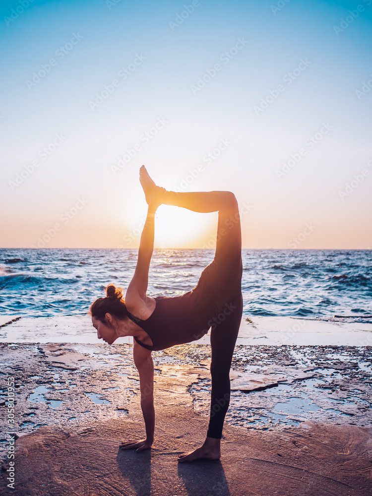 Young woman with curly hair in black costume practicing yoga at sunrise light, minimalist scene. Doing asana. Healthy lifestyle concept.
