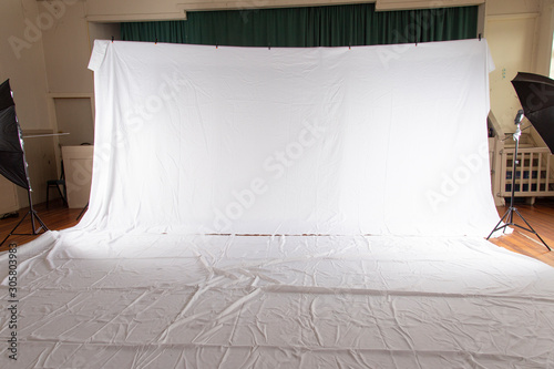 Pull back of set up for professional photography shoot. Volume portrait session. White muslin backdrop 6 metres wide by 3 meters long. Floor cover same size. Shows wrinkles and umbrellas on side. photo