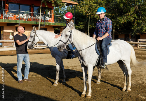 Woman and man with trainer riding horse at farm at summer day