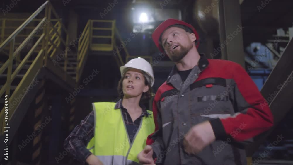 Waist-up tracking shot of Caucasian male and female manufacturing plant employees walking together through premises, looking around at machinery and discussing operational issues Waist-up tracking shot of Caucasian male and female manufacturing plant employees walking together through premises, looking around at machinery and discussing operational issues
