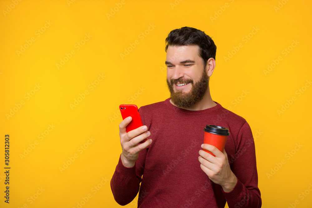 Bearded young man in red sweater is smiling and holding a smartphone and a cup of coffee on yellow background. Bearded young man in red sweater is smiling and holding a smartphone and a cup of coffee on yellow background.