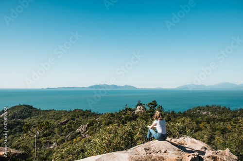 Young stylish independent girl travel the world, sitting on the rock and enjoying the view over the ocean and trees