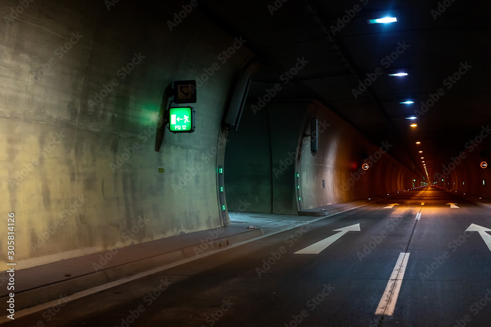 Automobile auto dark car tunnel with white arrows on asphalt showing way direction. Emergency exit sign with many lights. Empty underground vehicle road. Urban abstract city transportation background