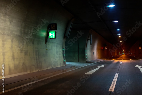 Automobile auto dark car tunnel with white arrows on asphalt showing way direction. Emergency exit sign with many lights. Empty underground vehicle road. Urban abstract city transportation background